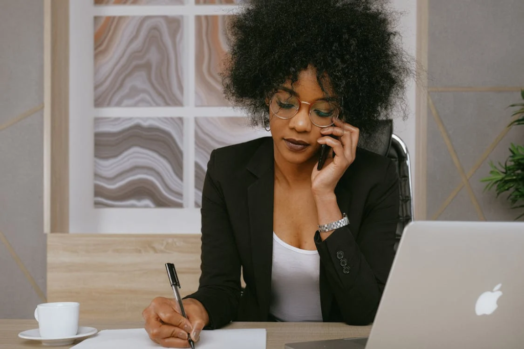 A woman working on her laptop and writing on paper while on the phone.